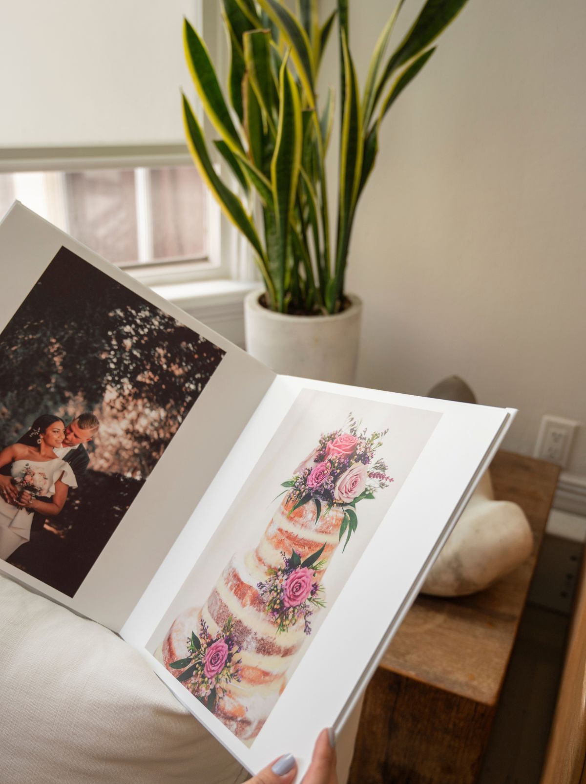 A hand holding open a layflat photo book, with a wedding portrait and a flowered cake on its pages