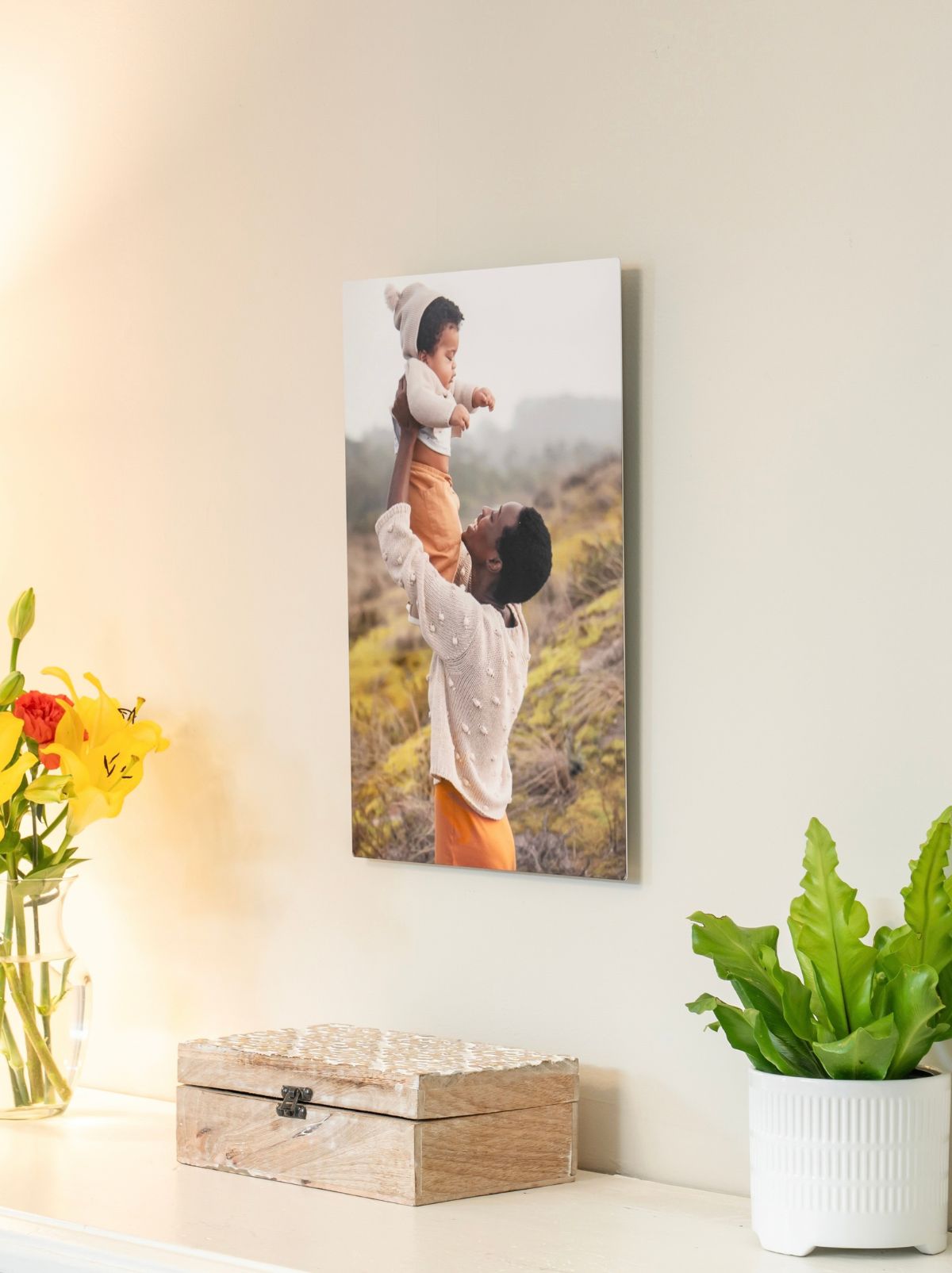 A metal print of a parent lifting a baby into the air, hanging above a shelf with flowers and a plant